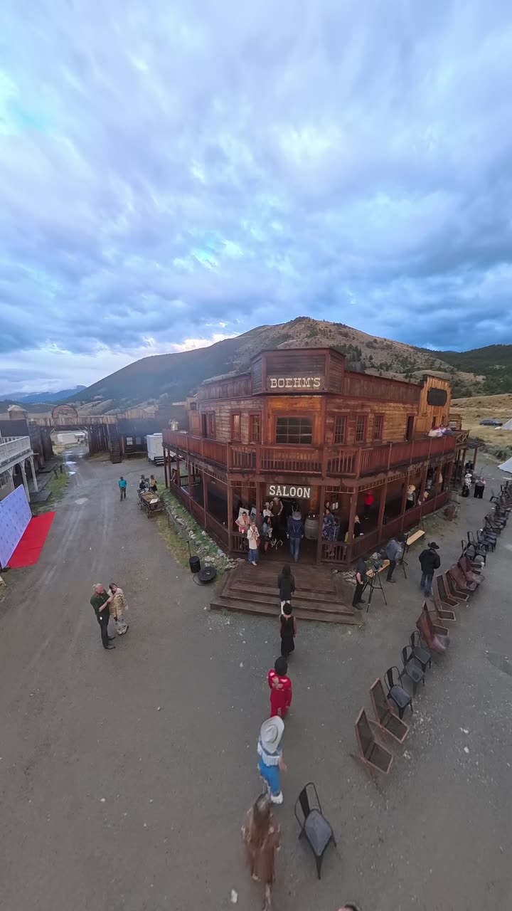 Looking down from above on a two-storey timber saloon with a balcony, people in period dress on the street below, rows of chairs set out, a mountain behind under heavy cloud.