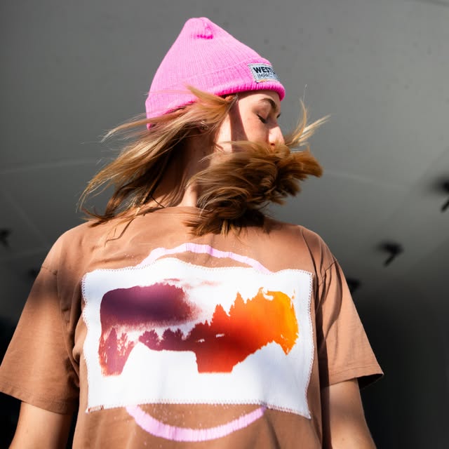 Person in a bright pink knit beanie and a tan T-shirt with a sunset-print graphic, hair mid-flip, photographed from below against a pale ceiling.