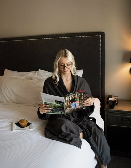 Person in a dark grey hotel robe sitting on a white bed reading a magazine, a muffin on a tray beside them, a warm lamp in the corner.