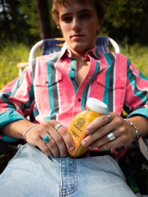 Person in a pink, teal and white striped shirt sitting in a camp chair among trees, holding a small bottle of orange juice, silver rings on their fingers.