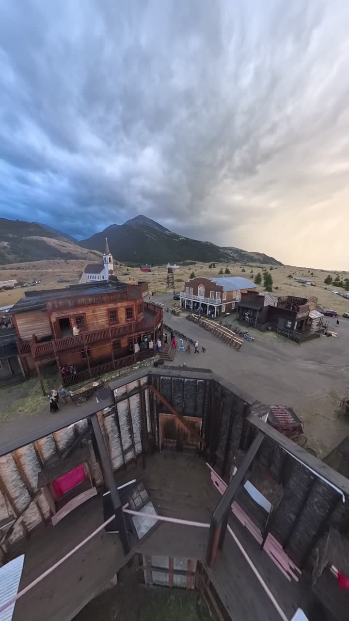 Looking straight down over a historic western town at last light: the open, roofless shell of a timber building fills the foreground, false-front timber buildings and a dirt main street beyond, a white timber church with a steeple further off, people gathered, vehicles parked at the edge of town, grassland and mountains under a broken sky going gold at the horizon.